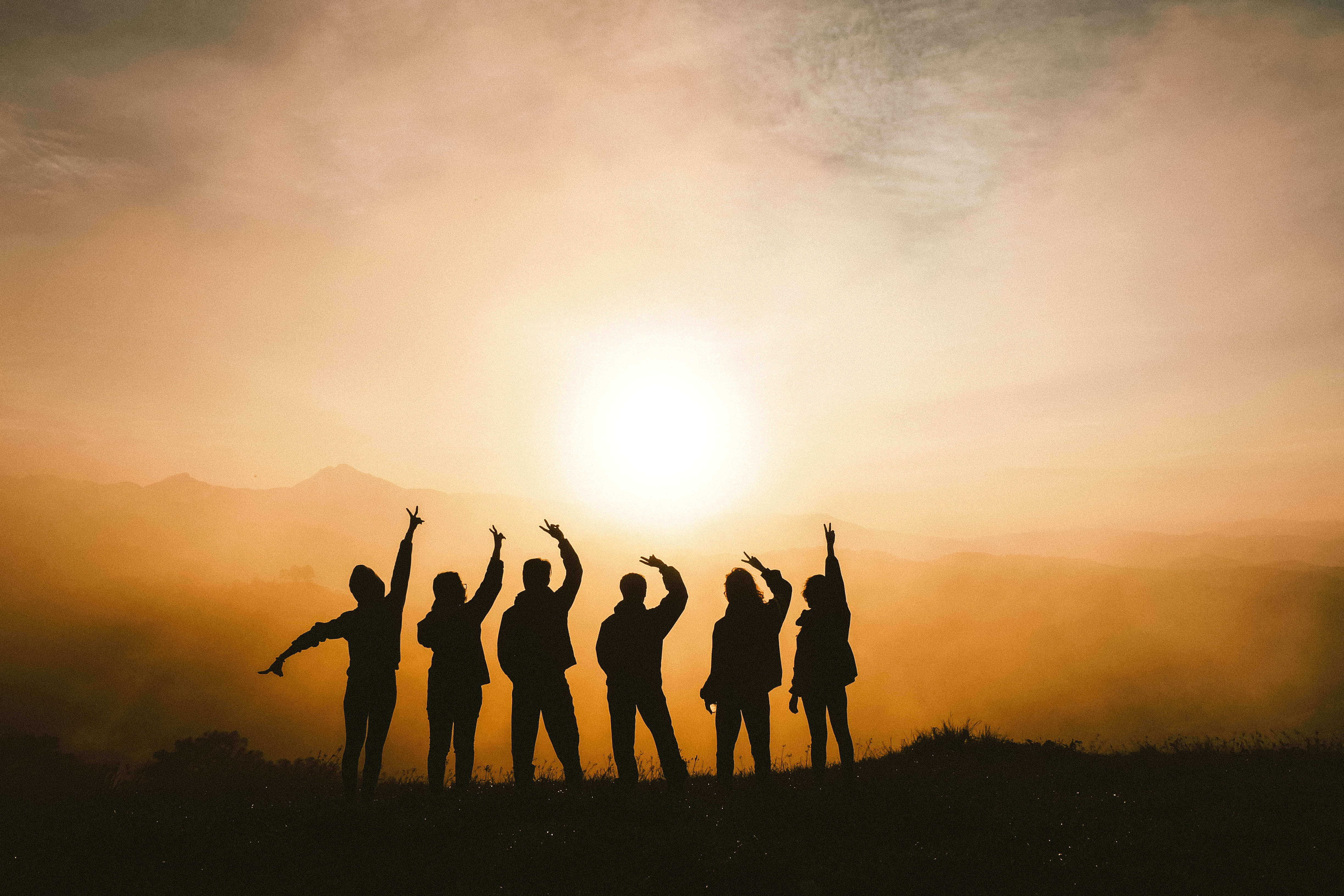 Silhouettes of several people against the sunset backdrop stretch their arms up in joy.
