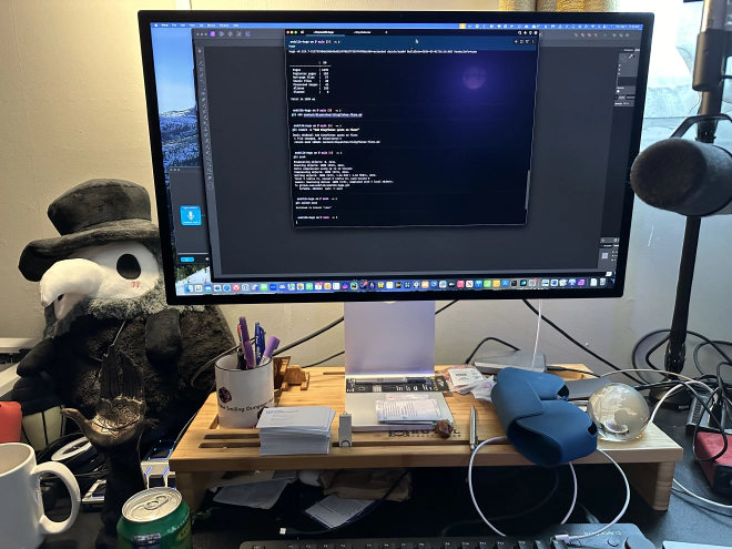 Photo of computer desktop highlighting a large monitor featuring a terminal, which sits upon a wood riser. There is a microphone on a boom arm, a cup of pens, various Numenera cards, a statue of a six-fingered hand, and a plushie plague doctor.
